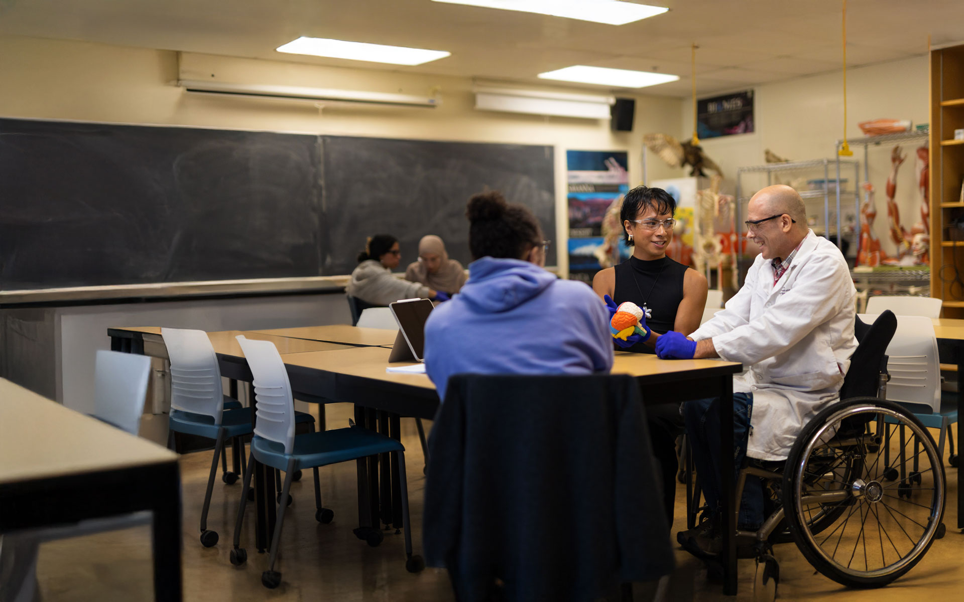 An instructor in a lab coat and wheelchair smiles while discussing an anatomical brain model with students at a table in a science classroom.