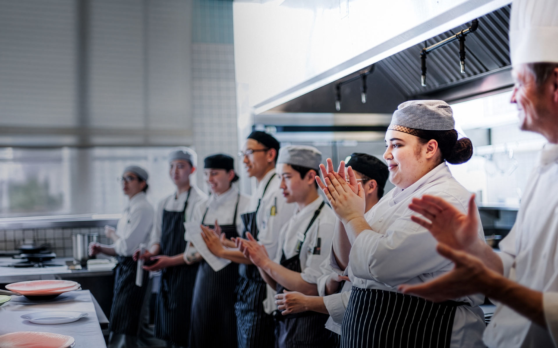 A line of culinary students and instructors in white chef uniforms and aprons stand in a commercial kitchen, smiling and applauding.