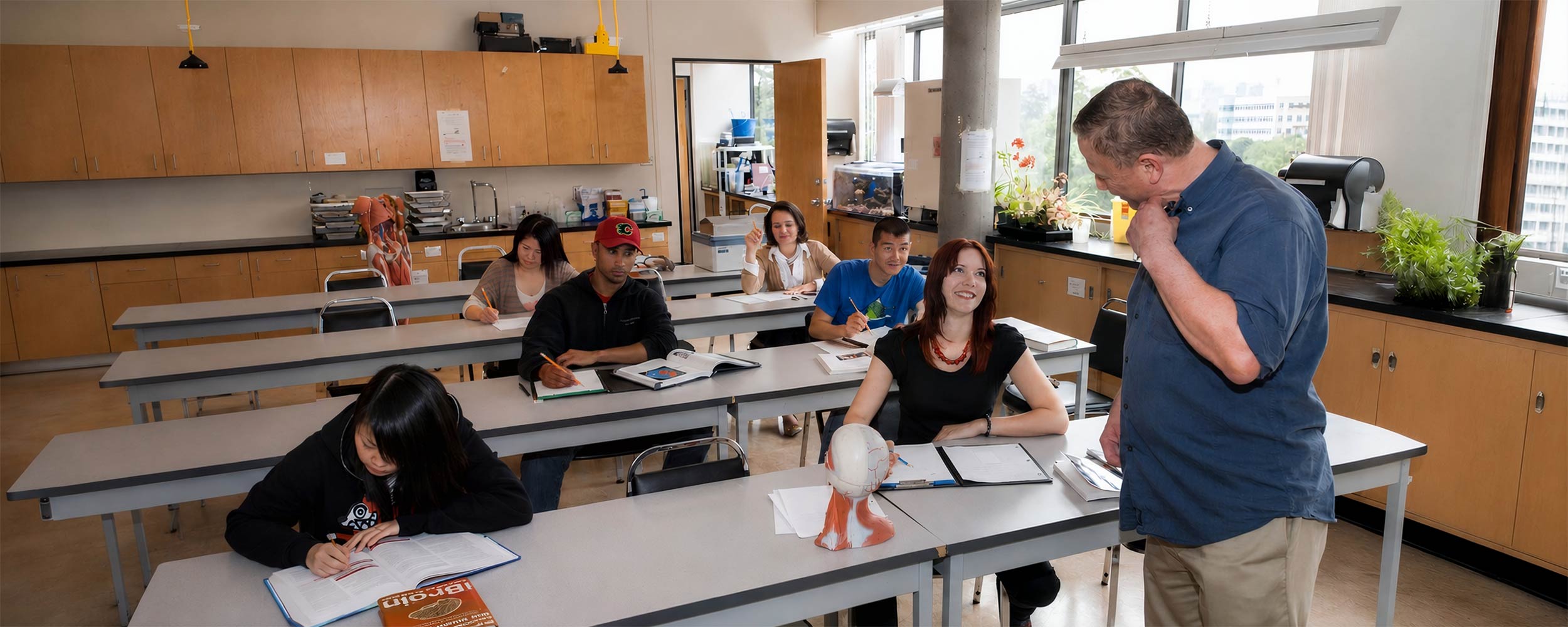 An instructor interacts with students seated at long lab desks during an academic upgrading science class.