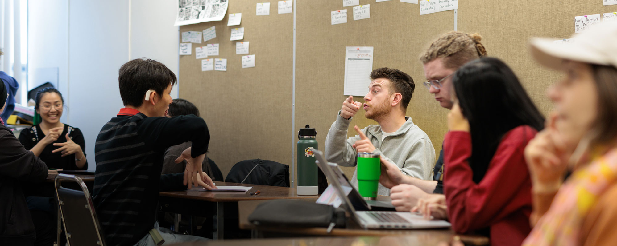 A group of students sit at desks and communicate using American Sign Language during a lesson in a classroom with a bulletin board in the background.