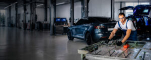 A mechanic in a blue jumpsuit and orange gloves works on the engine of a car in a garage setting.