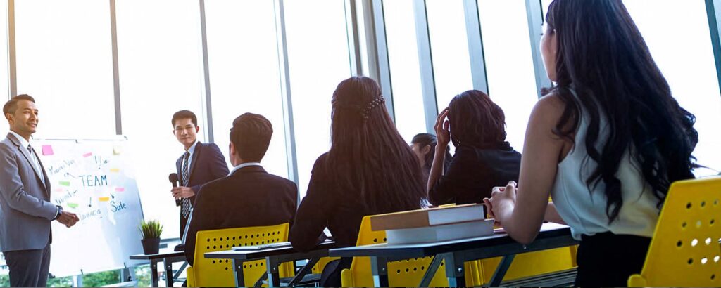 An instructor in a suit stands next to a whiteboard while presenting to business students seated in a brightly lit room with large windows.
