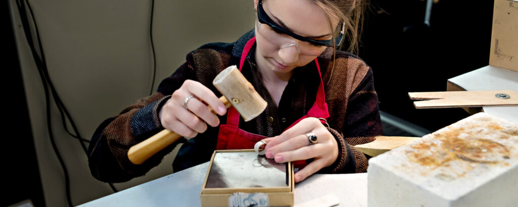 A jewellery student wearing safety glasses and a red apron uses a small wooden mallet to shape metal jewellery at a workbench.