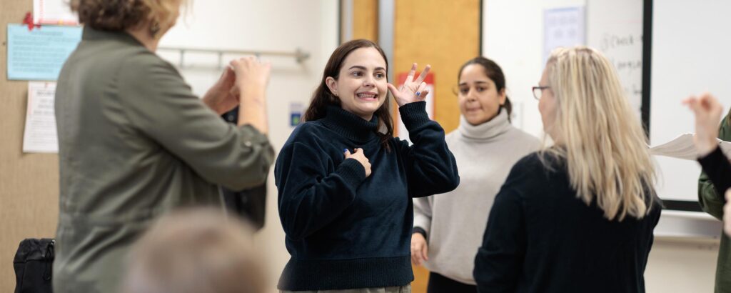 A group of people stand in a circle practicing sign language together in a classroom setting, with a smiling woman in a dark turtleneck actively demonstrating hand gestures.