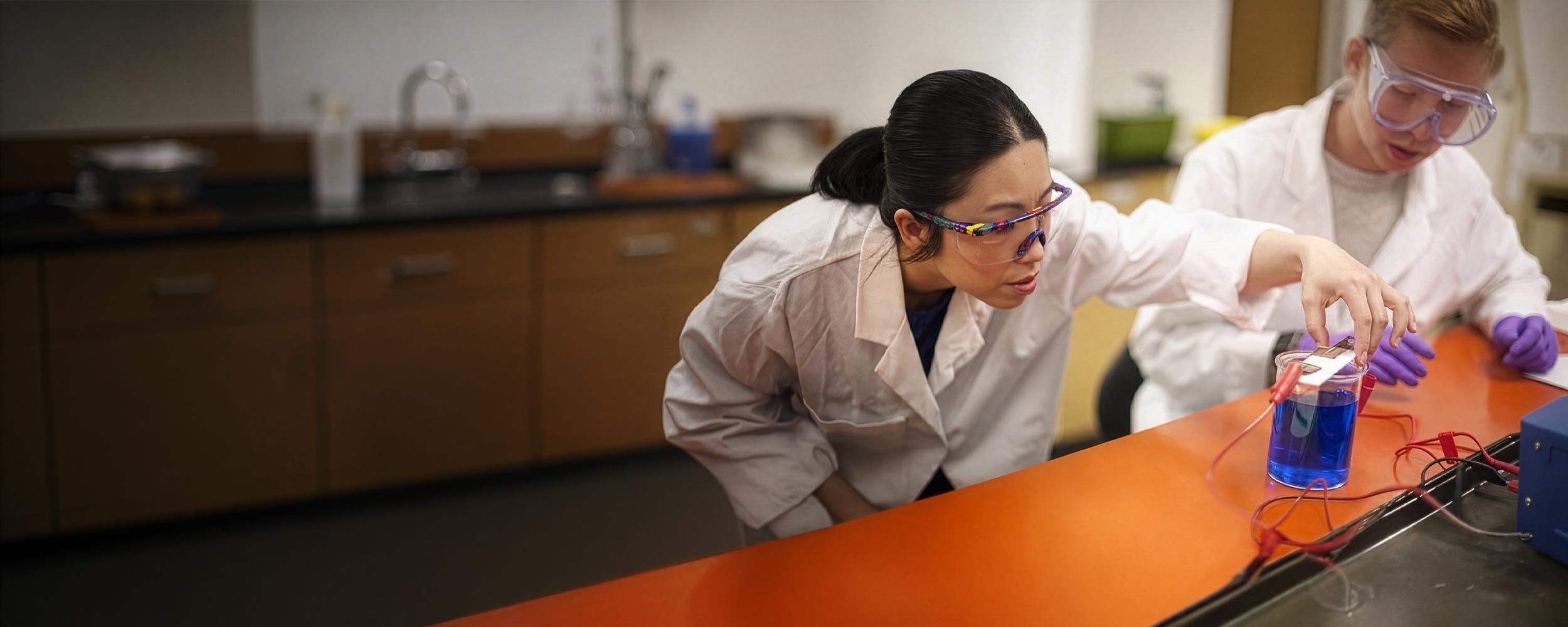 Two students in white lab coats and protective safety glasses conduct an chemistry or electrochemistry experiment, adjusting electrodes or leads connected to a beaker filled with a vivid blue liquid.