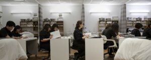 Esthetics students wearing black uniforms sit at individual desks studying coarse material inside a specialized spa training classroom equipped with treatment tables.